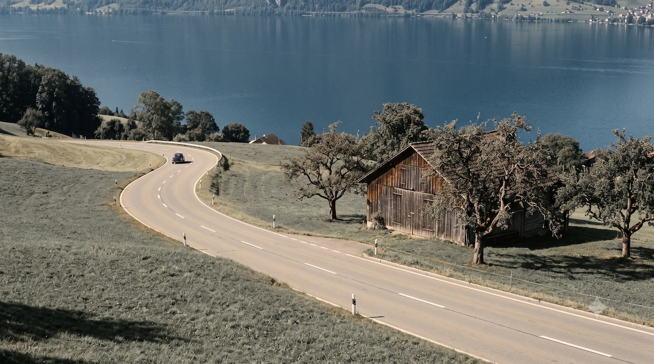 Eine Landstrasse durch Wiesen hoch über dem See, ein Wagen weit voraus.
